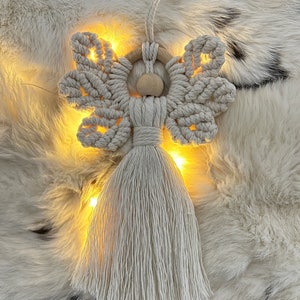 Macramé angel ornament with braided wings, a wooden bead head, and a long fringed tassel body, displayed on soft white fur with warm fairy lights glowing behind it.