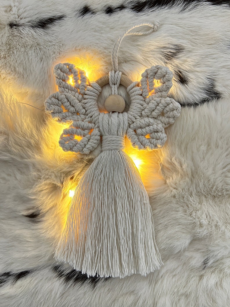 Macramé angel ornament with braided wings, a wooden bead head, and a long fringed tassel body, displayed on soft white fur with warm fairy lights glowing behind it.