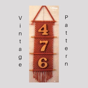 Puede incluir: Un tapiz de macramé rojo con números de madera 4, 7 y 6. Los números están tejidos en el diseño de macramé.