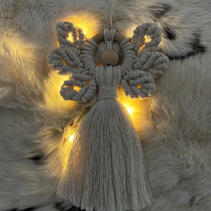 Macramé angel ornament with braided wings and a long tassel, shown on soft fur with warm fairy lights glowing behind it.
