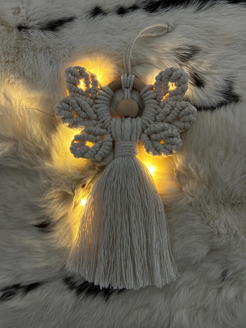 Macramé angel ornament with braided wings and a long tassel, shown on soft fur with warm fairy lights glowing behind it.