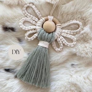 May include: A macrame ornament featuring a natural wooden bead head, cream-colored cord wings, and a sage green cord tassel body. The ornament includes a loop for hanging and a circular wooden ring frame. The text DIY is printed on the image.