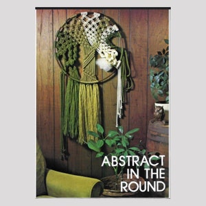 May include: A macrame wall hanging with green, white, and brown yarn. The hanging is in the shape of a tree and is attached to a wooden wall. The text "ABSTRACT IN THE ROUND" is printed below the hanging.