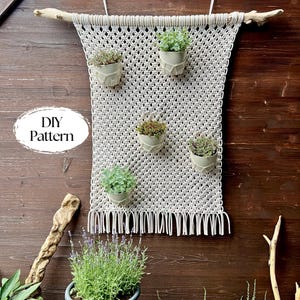 Peut inclure: Un porte-plantes en macramé avec cinq petites plantes en pot. Le macramé blanc cassé est suspendu à une branche de bois flotté. Le texte "DIY Pattern" est dans un ovale blanc à gauche. Le fond est un mur en bois foncé.