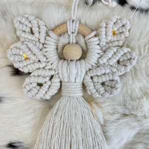 Handmade macramé angel ornament featuring textured wings, a wooden bead head, and a long tassel body, displayed on soft fur with warm lights in the background