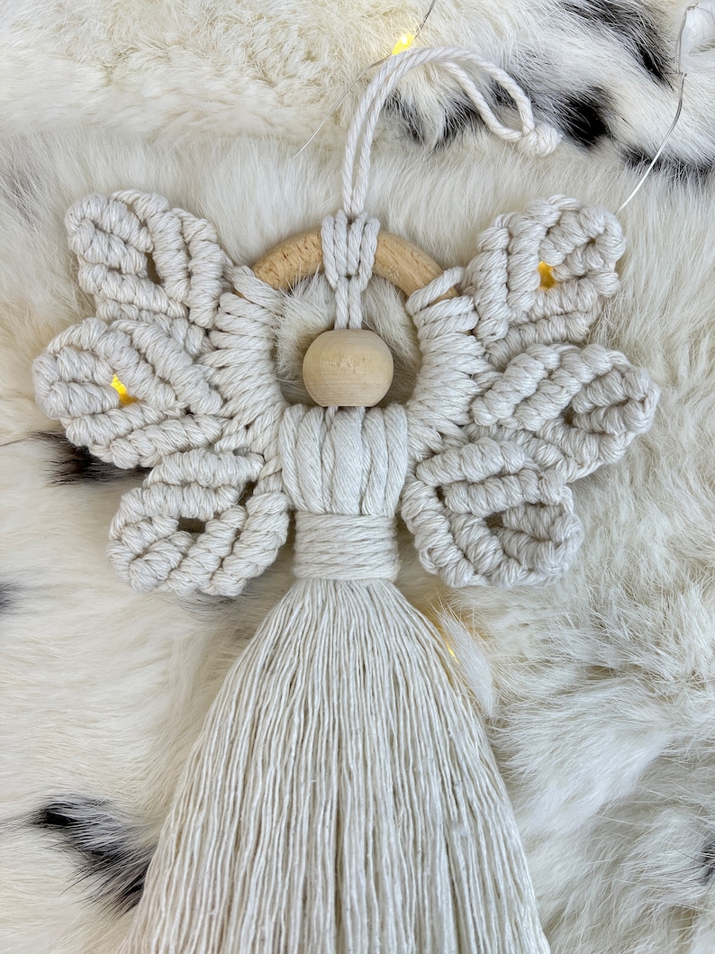 Handmade macramé angel ornament featuring textured wings, a wooden bead head, and a long tassel body, displayed on soft fur with warm lights in the background