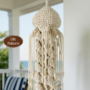 May include: A cream-colored macrame jellyfish wall hanging featuring a rounded top and long, knotted, dangling tentacles. A brown oval graphic on the left side contains the white text DIY Pattern. The piece is constructed from thick cord.