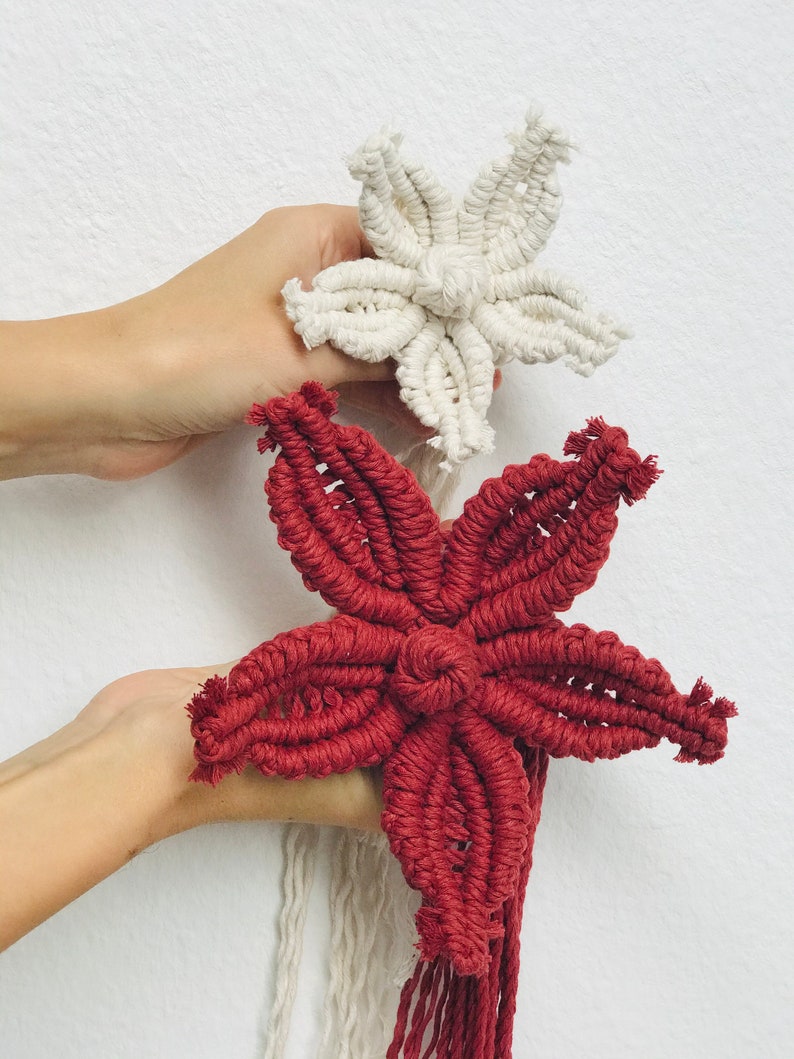 Puede incluir: Dos adornos de estrellas de macram&eacute;, uno blanco y otro rojo, colgando de una pared blanca.