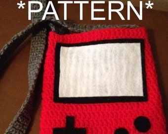 May include: Red crocheted bag with a black and white screen and buttons, resembling a handheld video game console. The bag has a gray crocheted strap. The text *PATTERN* appears above and below the bag.