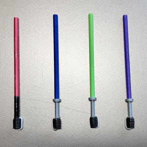 Peut inclure: Quatre sabres laser jouets, rouges, bleus, verts et violets. Chacun a une poignée argentée et une poignée noire. Les sabres laser sont disposés verticalement sur un fond gris clair.