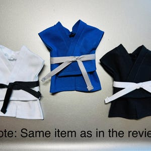 May include: Three miniature karate uniforms in white, blue, and black. Each uniform has a matching belt and is displayed on a silver surface. The image includes the text "Note: Same item as in the review."