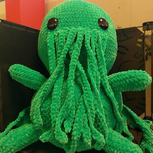 May include: A large, plush, green Cthulhu doll with large, dark eyes and multiple tentacles. The doll is made of a soft, textured material and has a round head and multiple arms. The tentacles hang down from the head.