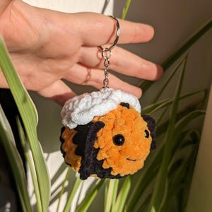 May include: A handmade, plush keychain in the shape of a bee. The bee is primarily orange with black accents and a white fluffy top. The keychain is attached to a silver metal ring.