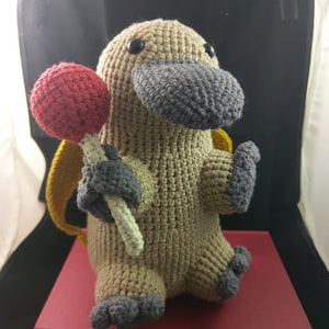 May include: A handmade crochet platypus doll in tan, gray, and red. The plush toy has a gray beak and feet, holding a red lollipop. It has a yellow backpack and is sitting on a red book.