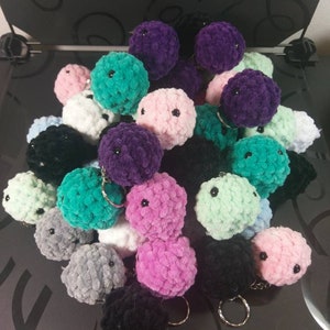 May include: A collection of colorful, plush ball keychains in various shades including purple, pink, teal, black, white, and gray. Each ball has a small, black eye and a metal keyring attached. The keychains are soft and fluffy.