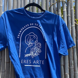 Könnte beinhalten: Ein königsblaues T-Shirt mit einem weißen Grafikdesign. Die Grafik zeigt eine Frau mit Blumen und dem Text "ENAMORATE DE TU EXISTENCIA ERES ARTE".