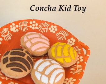 Può includere: Quattro conchas di peluche, un pane dolce messicano tradizionale, in una varietà di colori e design. Le conchas sono su un piatto con un motivo rosso e bianco. Concha Kid Toy è scritto sopra il piatto.