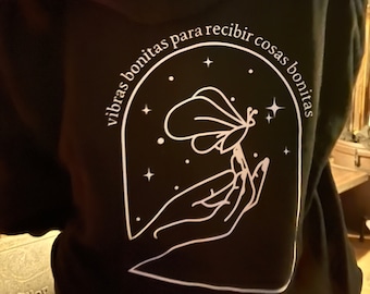 May include: Black hoodie with a white graphic of a hand holding a butterfly with the text "vibras bonitas para recibir cosas bonitas" in a circle around the graphic.