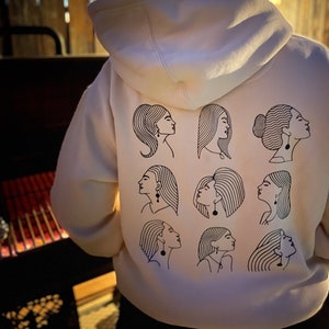 May include: White hoodie with a black line drawing of nine women's faces on the back. Each face has a unique hairstyle and is wearing earrings.