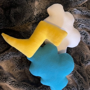 May include: Three plush pillows in the shapes of a cloud, a lightning bolt, and another cloud. The cloud pillows are white and blue, and the lightning bolt pillow is yellow.