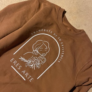 Könnte beinhalten: Ein braunes Sweatshirt mit einem weißen Grafikdesign, das ein Frauen Gesicht, Blumen und den Text "ENAMORATE DE TU EXISTENCIA ERES ARTE" zeigt.