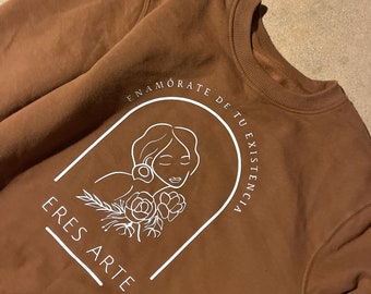 May include: A brown sweatshirt with a white graphic design featuring a woman's face, flowers, and the text "ENAMORATE DE TU EXISTENCIA ERES ARTE".