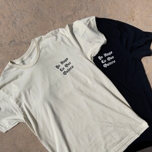 May include: Two t-shirts, one white and one black, with the same text in black lettering: "Yo Hago Lo Que Quiero".
