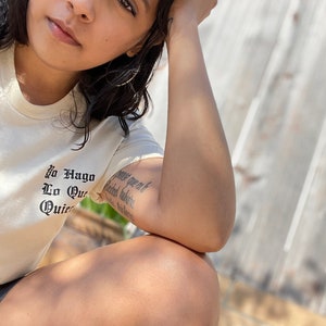 May include: A person wearing a white t-shirt with the text "Yo Hago Lo Que Quiero" in black lettering. The person has a tattoo on their arm.