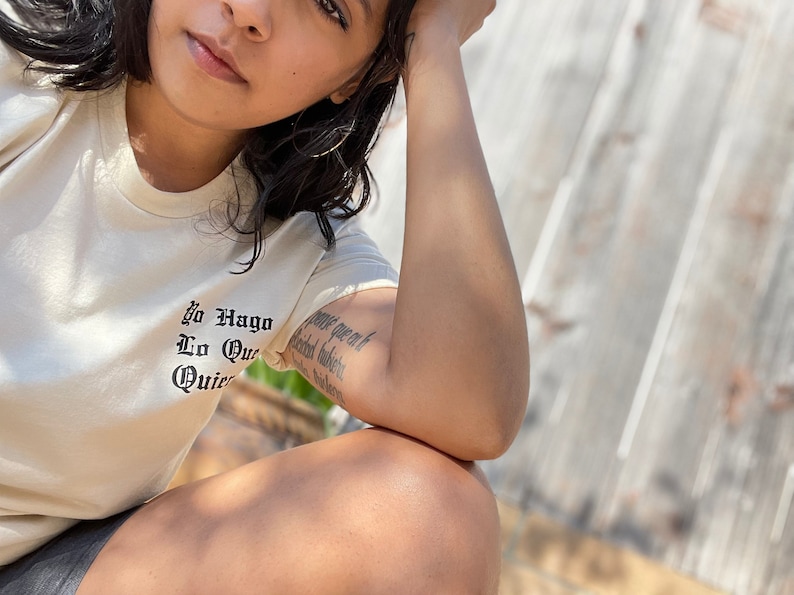 May include: A person wearing a white t-shirt with the text "Yo Hago Lo Que Quiero" in black lettering. The person has a tattoo on their arm.