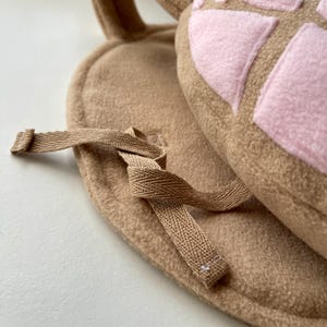 May include: A tan-colored, plush baby hat with attached ears and a matching bib. The hat features a pink, patterned design on the top. A tan fabric tie is attached to the bib. The set is made of soft, textured material.