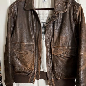 Puede incluir: Una chaqueta bomber de cuero marrón con cierre de cremallera y dos bolsillos grandes. La chaqueta tiene un cuello, puños y cintura de punto de canalé. La chaqueta tiene una etiqueta en el interior que dice "Black bellies of anti-navy dome".