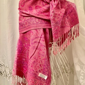 May include: A pink paisley scarf with fringe. The scarf is draped over a white wire mannequin. The scarf has a detailed pattern with shades of pink and purple. The fringe is a matching pink color. The scarf is made of a soft material.
