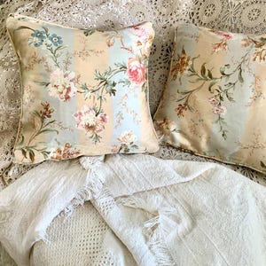 May include: Two square throw pillows with floral patterns. The pillows feature a cream, light blue, and peach striped background with pink, blue, and cream-colored flowers and green leaves. The pillows are placed on a white, textured blanket.