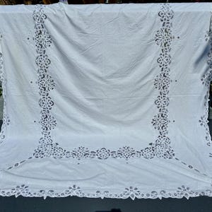 May include: A white rectangular tablecloth featuring an intricate cutwork lace border and a matching lace panel inset. The fabric is smooth and opaque, with detailed floral and geometric embroidery patterns integrated into the lace sections.