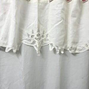 May include: White linen shower curtain with a decorative lace trim along the bottom edge. The curtain is hung with metal hooks adorned with pink flower designs. The curtain is made of a lightweight fabric.
