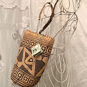 May include: A woven basket bag with a geometric pattern in shades of brown and tan. The bag has a braided handle and a tag attached. The bag is displayed on a white wire mannequin against a backdrop of white lace.