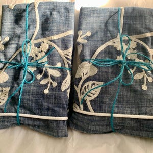 May include: Two folded denim-look fabric items with white floral embroidery are tied with turquoise twine. The fabric has a white rope trim. The items are on a white surface. The overall theme is home decor.