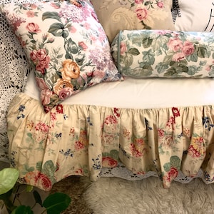 May include: A beige bed skirt with a floral pattern featuring pink, red, and blue flowers. The skirt has a ruffled edge and is decorated with a white lace trim.
