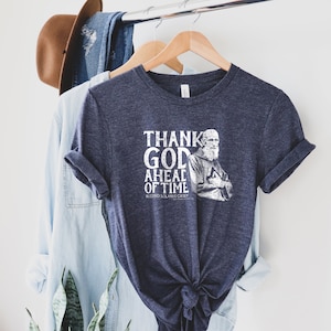 Puede incluir: Una camiseta azul marino con un gráfico blanco de un hombre con el texto "Thank God Ahead of Time" y "Blessed Solanus Casey".