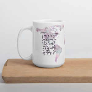 May include: White ceramic mug featuring a watercolor world map design in shades of pink and purple. The mug has the text "There is still some good in this world & it is worth fighting for". The mug is on a wooden surface.
