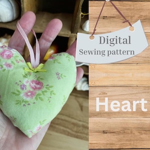 Può includere: Un modello di cucito digitale per un cuscino a forma di cuore. Il cuscino è verde con una stampa floreale e ha un nastro rosa legato intorno. Il testo "Modello di cucito digitale" e "Cuore" è visibile nell'immagine.