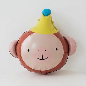 May include: A brown and pink monkey balloon with a yellow crown and a blue top. The balloon has a smiling face with black eyes and a small nose.