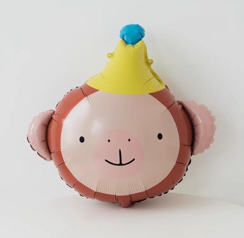 May include: A brown and pink monkey balloon with a yellow crown and a blue top. The balloon has a smiling face with black eyes and a small nose.