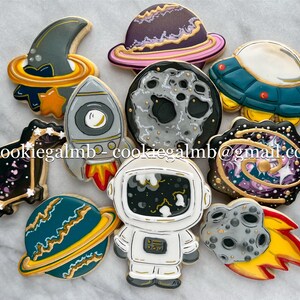 May include: A set of nine space-themed sugar cookies decorated with colorful icing. The cookies feature a rocket, an astronaut, a crescent moon, a planet, a UFO, a galaxy, and three different moons.