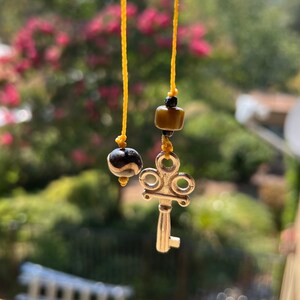 Puede incluir: Un dije de llave plateado con un diseño decorativo cuelga de una cuerda amarilla con cuentas negras y marrones. El dije está colgando frente a un fondo borroso de árboles verdes y flores rosas.
