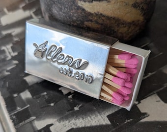 May include: A rectangular silver-toned metal matchbox case featuring the embossed text Allens est. 2010 on the front. The case is partially open, revealing a collection of wooden matches with bright pink tips tucked inside the white inner tray.