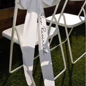 Op de afbeelding: Twee witte klapstoelen met een witte stoffen strik en een lint. Op het lint staat in zwart schrift "Row Reserved". De stoelen staan op een groene ondergrond, waarschijnlijk gras, en zijn opgesteld voor een evenement.