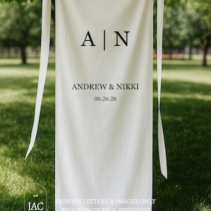 May include: A white banner with black text, hanging from a tree branch. The banner reads "A | N" above "ANDREW & NIKKI" and the date "06.26.26". The bottom of the banner is gathered. The text "IRON ON LETTERS & IMAGES ONLY NO LINEN OR SIGN INCLUDED" is also visible.