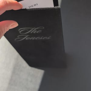 May include: A black rectangular menu cover with a soft texture, featuring the text "The Joneses" in a silver script font. The cover has a gold-coloured metal corner protector and reveals a white inner page with the text "DEBI" at the top.
