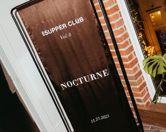 Pode incluir: Um banner castanho com o texto branco "SUPPER CLUB Vol. 8" e "NOCTURNE". A data "11.21.2025" também é exibida. O banner é suportado por uma estrutura de metal preta, contra uma parede de tijolos.